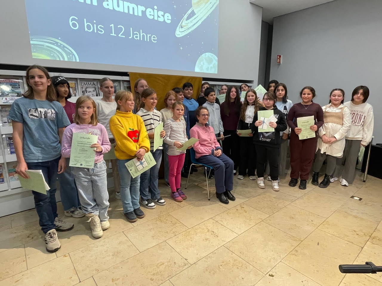 Jugendpreis 2025 6 bis 12 auf der linken Seite die Teilnehmerinnen und Teilnehmer am Wettbewerb „Weltraumreise“ in der Mitte sitzt die Stifterin der Arno Reinfrank  Jugendpreises Frau Jeanette Koch -Reinfrank und auf der rechten Seite sehen wir die anwesenden Teilnehmerinnen und Teilnehmer aus der Kategorie Bildergeschichte ( F&ouml;rderklasse im Fach Deutsch)